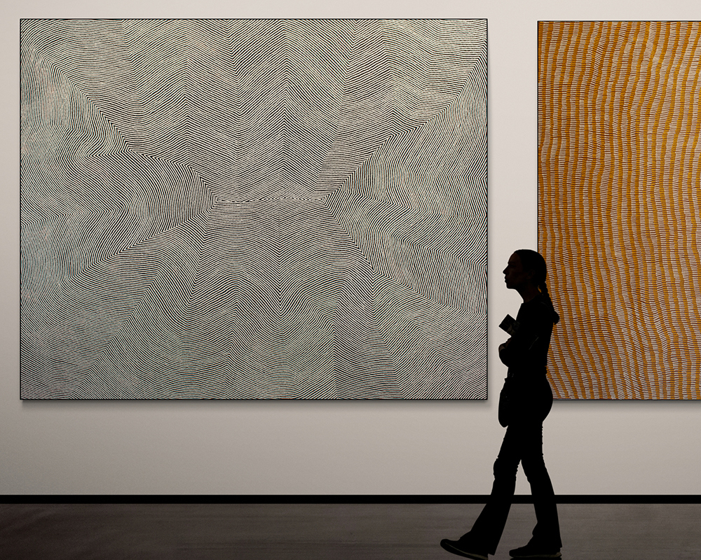 Pintupi Nine Artworks hanging in a gallery, with a silhouetted woman walking past